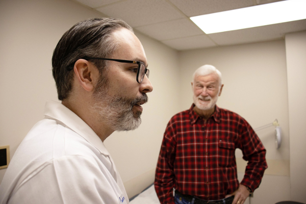 A physician in a white coat talks with an older male patient in an exam room. The patient, wearing a red plaid shirt, stands smiling as they speak.